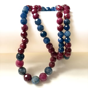 Pode incluir: Um colar composto por contas redondas facetadas em cores vermelho profundo e azul escuro alternadas. As contas estão enfiadas num fio contínuo, exibindo uma textura de superfície reflexiva e multifacetada ao longo de toda a peça.