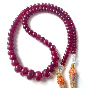 Peut inclure: Un collier composé de perles circulaires polies d'un rouge profond disposées en boucle. Les perles varient légèrement en taille, avec les plus grandes vers le bas. Les extrémités sont finies avec des embouts argentés, du fil doré et du fil orange.