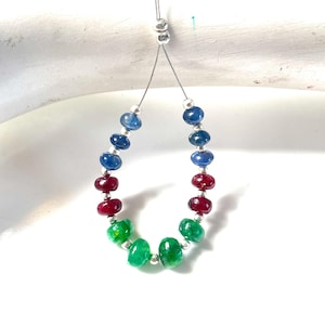 Puede incluir: Un hilo de pequeñas cuentas de piedras preciosas lisas y redondas en tonos azul, rojo y verde, separadas por diminutos espaciadores de metal plateado. Las cuentas están dispuestas en forma de U sobre un hilo fino, con tres azules, dos rojas.