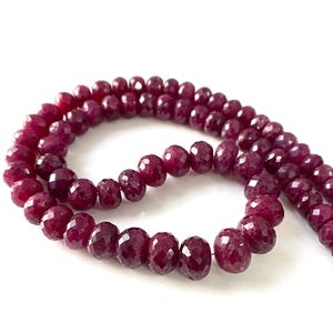 Peut inclure: Un collier de perles en pierre précieuse rouge foncé, circulaires et facettées, disposées en une formation courbe et lâche. Chaque perle présente de multiples surfaces polies qui reflètent la lumière, créant un aspect texturé le long du fil.