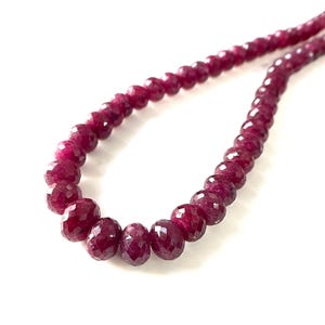 Può includere: Un filo di perline di rubino sfaccettate di un rosso magenta intenso disposte in linea curva. Le perline sono rotonde e presentano una superficie lucida e riflettente con molteplici piccole sfaccettature geometriche che catturano la luce.