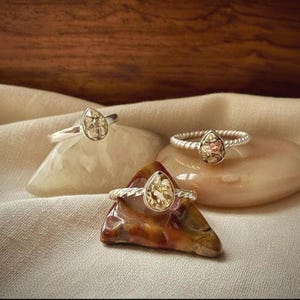 May include: Three silver rings featuring teardrop-shaped settings with gold-toned flecked inlay. One ring has a smooth band, while the other two feature twisted, rope-like bands. Each ring is displayed resting on a smooth, polished stone surface.