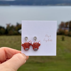 May include: A pair of dangle earrings featuring brown Highland cow charms with white horns and grey noses. Each earring is attached to a small, clear, round rhinestone stud. The earrings are displayed on a white card with the text Clay & Cozy Knits.
