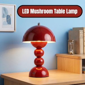 Puede incluir: Lámpara de mesa con forma de seta LED roja con una pantalla redondeada y una base de esferas apiladas. El texto "LED Mushroom Table Lamp" está en una pancarta roja. La lámpara tiene un puerto USB.