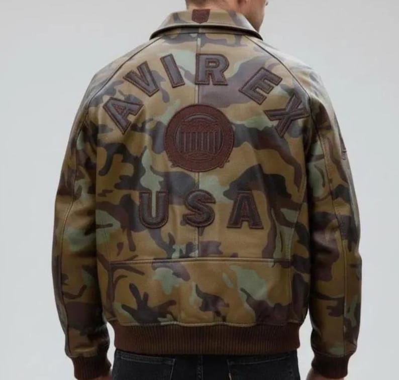 May include: A camouflage-patterned leather jacket with the word "AVIREX" arched across the shoulders and "USA" below. The jacket features a brown collar, cuffs, and hem, and a circular emblem. The overall color palette includes shades of brown, green, and black.