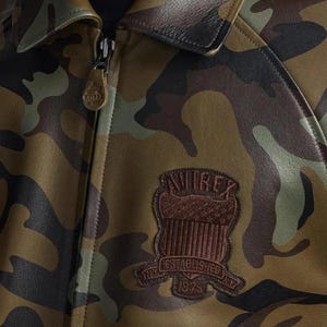 May include: Camouflage-patterned leather jacket with a brown leather collar and a zipper closure. The jacket features a brown patch with the text "AVIREX" and "ESTABLISHED 1975 NY NY". The jacket's color palette includes shades of green, brown, and tan.