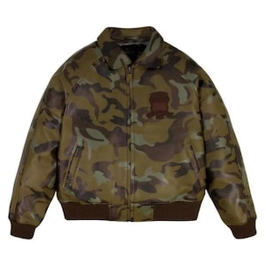 May include: A camouflage-patterned jacket in shades of green, brown, and tan. The jacket features a collared neckline, a front zipper, and a brown ribbed hem and cuffs. A small emblem is visible on the upper left chest.