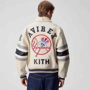 Può includere: Giacca in pelle color crema con le scritte "AVIREX" e "KITH" in blu navy. La schiena presenta un logo a tema baseball con "Yankees" in rosso e bianco. Le maniche hanno strisce blu navy e grigie. Una giacca elegante per gli appassionati di sport.
