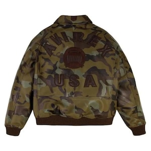May include: Camouflage-patterned leather jacket with the word "AVIREX" arched across the back, and "USA" below. The jacket has a brown ribbed hem and cuffs. The camouflage design features shades of green, brown, and black.
