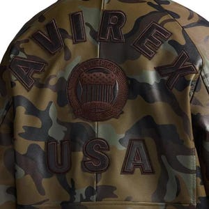 May include: Camouflage-patterned leather jacket with the word "AVIREX" and "USA" in dark brown lettering. A circular patch with text and an American flag design is centered on the back of the jacket.