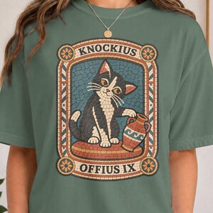 May include: A sage green t-shirt featuring a graphic print of a black and white cat sitting on a cushion next to a terracotta-coloured vase. The design is framed by a decorative border with the text KNOCKIUS at the top and OFFIUS IX at the bottom.