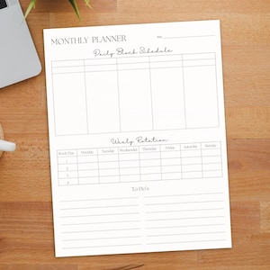 May include: A white monthly planner with the text "Monthly Planner" and "Daily Block Schedule" is on a wooden surface. The planner includes sections for daily and weekly scheduling. A laptop and a cup of coffee are in the background.