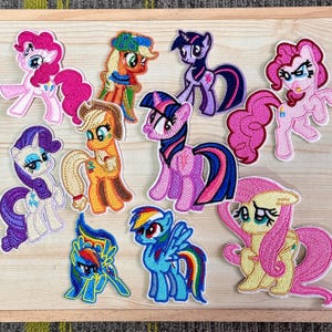 Könnte beinhalten: Eine Sammlung farbenfroher, gestickter Aufnäher mit verschiedenen My Little Pony-Charakteren. Die Aufnäher zeigen Ponys in Rosa, Lila, Blau, Gelb und Orange, mit detaillierten Gesichtsausdrücken und Mähnen. Diese Aufnäher eignen sich ideal zum Dekorieren von Kleidung oder Accessoires.