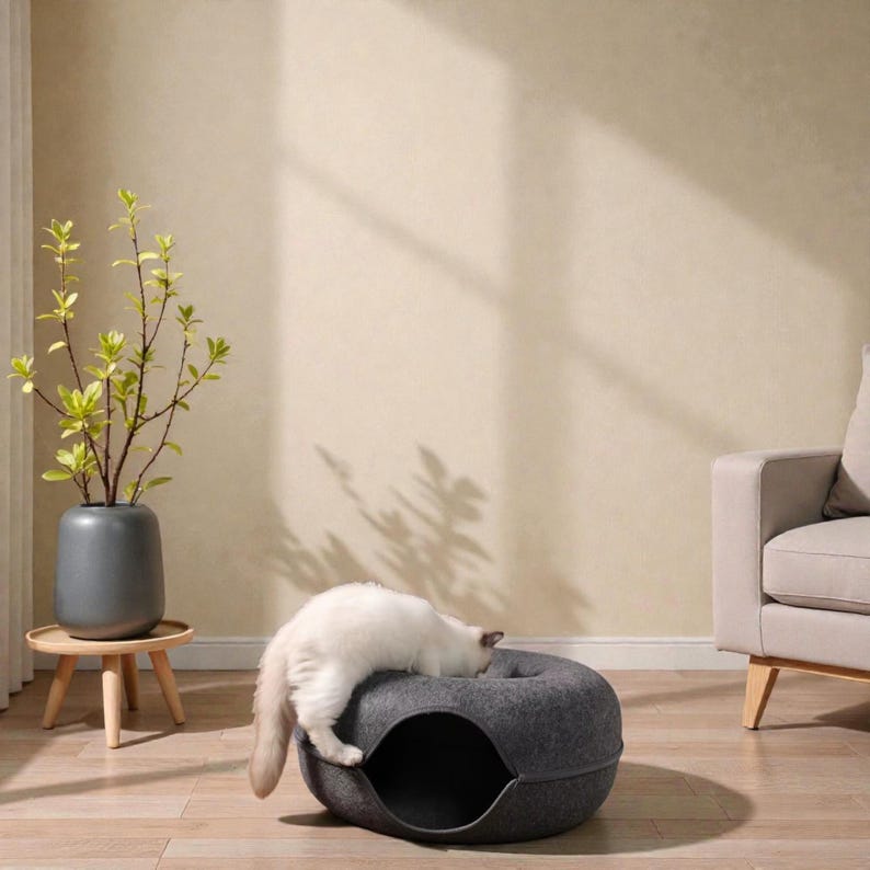 May include: A gray cat bed with a circular design and a rounded opening. A white cat is resting on top of the bed. The bed is made of a soft, felt-like material. A small, wooden side table with a gray vase and green plant is in the background.