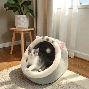 Può includere: Un letto per gatti grigio con finiture bianche e rosa e un giocattolo a pompon bianco pendente. Un gatto grigio e bianco è all'interno del letto, che guarda la fotocamera. Il letto per gatti è su un tappeto colorato.