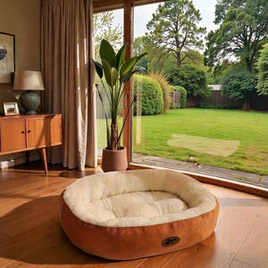 May include: A circular pet bed with a brown outer and a cream-coloured, plush inner. The bed sits on a wooden floor, in front of a large window overlooking a green lawn. A potted plant is next to the bed.