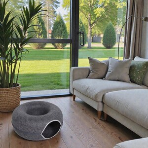 May include: A gray, donut-shaped pet bed with a white-trimmed entrance sits on a wooden floor. The bed is in a living room with a large window overlooking a green lawn and trees. A sofa with neutral-colored cushions is also visible.