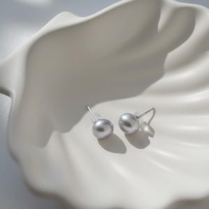 Peut inclure: Une paire de boucles d'oreilles à clous en perles grises avec des tiges en argent, posées sur un plat blanc en forme de coquillage. Les boucles d'oreilles sont rondes et ont un éclat subtil. Le plat a une finition lisse et brillante.