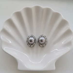 Könnte beinhalten: Ein Paar graue Perlenohrstecker mit silberfarbenem, eingelegtem Rand. Die Ohrringe werden auf einer weißen, muschelförmigen Schale präsentiert. Klassischer Schmuck.