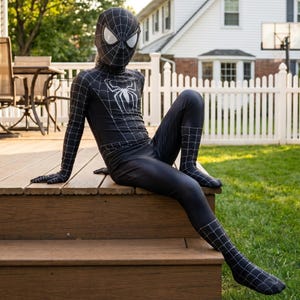 Op de afbeelding: Een zwart Spider-Man-kostuum met een wit spinnenembleem en witte webdetails. Het full-body pak bevat een bijpassend masker met witte ooglenzen. Het kostuum wordt gedragen door een persoon die op een houten terras zit.