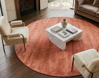 May include: A living room scene featuring a large, round, coral-colored rug. Two cream-colored armchairs with gold frames and a marble coffee table are arranged on the rug. A fireplace and a sofa are visible in the background.