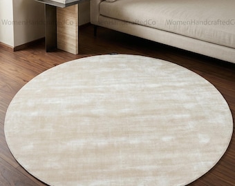 May include: A large, round, neutral-toned rug with a soft, textured surface. The rug is placed on a dark wood floor, and the overall aesthetic is minimalist and modern. The rug's diameter is approximately 8 feet.
