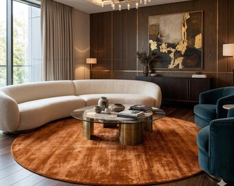 May include: A modern living room featuring a large, curved cream-colored sofa, two teal armchairs, and a round, rust-colored rug. A glass-topped coffee table with brass base sits in the center. The room has dark wood paneling and a large abstract painting.