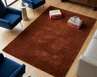 May include: A large, rectangular, rust-colored rug dominates the scene, complemented by two navy blue armchairs and a gray sofa. Two small marble side tables with gold legs and a wooden cabinet add to the decor. Books and an ashtray are placed on the rug.