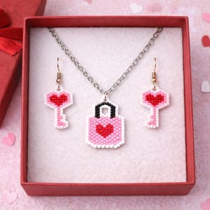 May include: A Valentine's Day jewelry set in a red gift box. The set includes a necklace with a lock pendant and earrings shaped like keys, all with pink and red heart designs. The box is next to a red gift box with a bow.