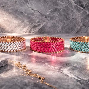 Op de afbeelding: Drie brede armbanden met geweven ontwerpen. Eén is wit, één is felroze en de derde is turquoise. Elke armband heeft een goudkleurige sluiting en afwerking. De armbanden worden gepresenteerd op een grijze, getextureerde ondergrond.