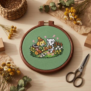 Peut inclure: Un cercle à broder au point de croix avec une scène de dessin animé de deux personnages sur fond vert. Le cercle en bois encadre le motif, qui comprend des fleurs et une clôture. Des fleurs séchées et un petit panier entourent la scène.