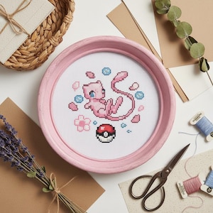 May include: A pink cross-stitch of the Pokémon Mew inside a round, pink frame. The design includes a Poké Ball, flowers, and blue bubbles. The frame is surrounded by craft supplies, including scissors, thread, and dried lavender.