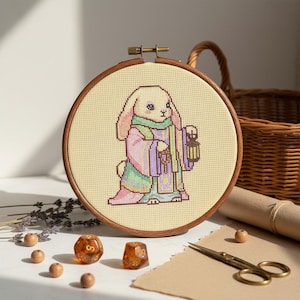 Pode incluir: Uma bordado em ponto cruz de um personagem coelho em uma túnica, segurando uma lanterna. A obra de arte está em um bastidor de bordado de madeira. O coelho está vestindo uma túnica com tons de rosa, verde e roxo. Dados e tesouras em primeiro plano.