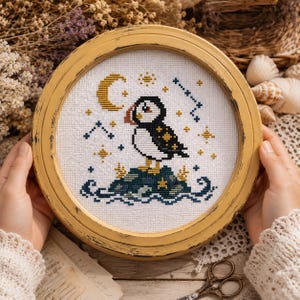 Puede incluir: Un marco redondo de madera color amarillo mostaza desgastado que contiene un bordado de punto de cruz de un frailecillo sobre una roca rodeada de agua. El diseño incluye una luna creciente amarilla, constelaciones azul oscuro y estrellas.