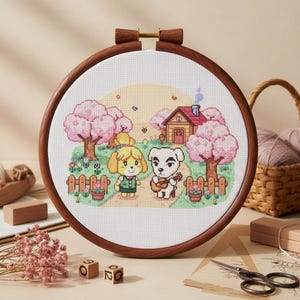 以下が含まれることがあります： 2匹の漫画のキャラクター、黄色の犬と白い犬が家の前の小道を歩いているクロスステッチ刺繍。シーンは木のフープで囲まれ、ピンクの木と小さな柵が含まれています。