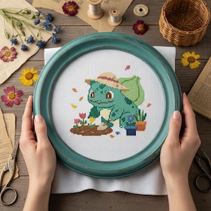 Op de afbeelding: Een kruissteekborduurwerk met een groen Bulbasaur-personage met een strohoed, die een tuin met bloemen verzorgt. Het kunstwerk is geplaatst in een ronde, blauwgroene lijst. De achtergrond bevat gedroogde bloemen en vintage items.