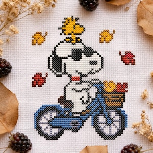 Può includere: Punto croce di Snoopy in bicicletta blu con Woodstock sopra. Il cestino è pieno di foglie autunnali. Lo sfondo include foglie autunnali e more.