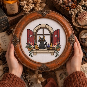 Peut inclure: Une broderie au point de croix circulaire encadrée dans un cadre en bois orange-brun vieilli avec des ornements métalliques. Le motif représente un chat noir assis dans une fenêtre avec des volets rouges, un balai et des fleurs en pot.