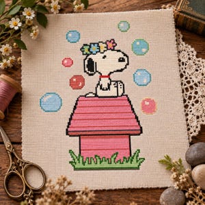 May include: A cross-stitch embroidery on beige fabric featuring a white dog with black ears wearing a colorful flower crown, sitting atop a pink doghouse. The design includes floating blue and pink bubbles and green grass at the base.