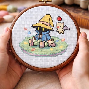 May include: A cross-stitch embroidery in a wooden hoop depicts a character with a yellow hat and a small, winged creature. The design is set on a light blue background with a green and pink grassy area. The hoop is held by two hands.