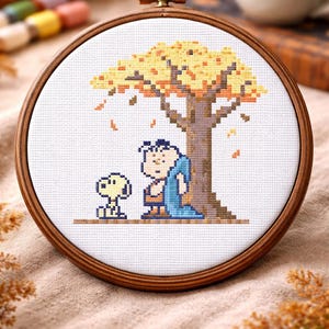 May include: Cross-stitch embroidery in a wooden hoop. The design features a cartoon of a person and a dog under a tree with yellow and orange leaves. The person is wearing blue and brown clothing. The dog is yellow. A cup of coffee is in the background.