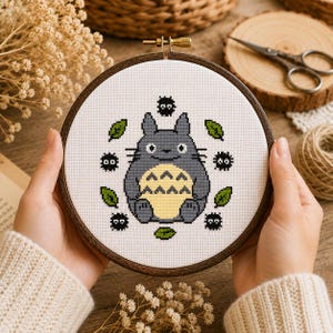 Può includere: Un telaio da ricamo a punto croce con un personaggio di Totoro grigio con la pancia gialla, circondato da foglie verdi e folletti neri della fuliggine. Il ricamo è in un telaio di legno.