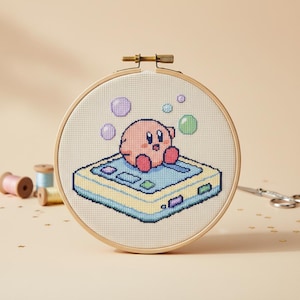 May include: A cross-stitch embroidery hoop featuring a pink character sitting on a light blue and yellow game console, with purple and green bubbles. The embroidery is set in a wooden hoop.