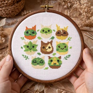 May include: A cross-stitch embroidery hoop featuring a circular design of stylized cat faces in various colors, including green, orange, and brown. The cats are surrounded by green leaves, all stitched on white fabric. The wooden hoop is held by hands.
