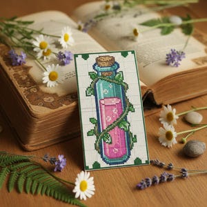 Könnte beinhalten: Ein Kreuzstich-Lesezeichen mit einem Fläschchen-Design. Die Flasche ist mit rosa Flüssigkeit gefüllt und mit grünen Ranken umwickelt. Das Lesezeichen hat einen grünen Rand und einen Korken. Gänseblümchen und ein offenes Buch sind im Hintergrund.
