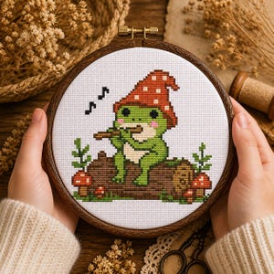 Könnte beinhalten: Ein Kreuzstich-Stickrahmen mit einer grünen Froschfigur, die einen roten Pilzhut trägt und auf einem Baumstamm Flöte spielt. Das Design umfasst Noten, Pilze und Grünzeug, alles vor einem weißen Hintergrund in einem Holzrahmen.