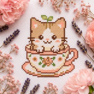 May include: Cross-stitch of a cute cat in a teacup. The cat is brown and white with a green leaf on its head. The teacup is decorated with pink flowers. The background is white fabric with pink flowers and lavender sprigs.