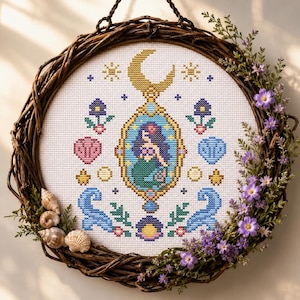 May include: A cross-stitch embroidery featuring a central mermaid inside an ornate gold frame topped with a crescent moon. The design includes blue waves, pink and blue seashells, small flowers, and stars, all mounted within a circular twig wreath.