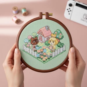 Puede incluir: Bordado de punto de cruz en un aro de madera con personajes de Animal Crossing. El diseño incluye una casa, flores y personajes regando plantas. El aro se sostiene frente a un fondo rosa.