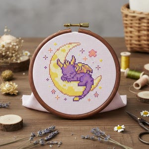 Op de afbeelding: Een borduurring met kruissteek met een paarse draak die slaapt op een gele halve maan. Het ontwerp is geplaatst op een lichtroze stof, omlijst door een houten ring. Decoratieve elementen zijn sterren en bloemmotieven.